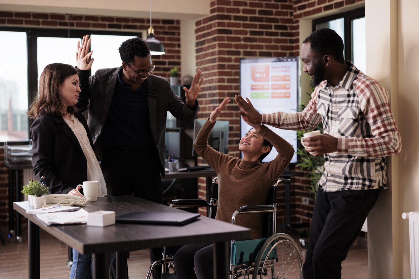 Multiethnic group of people having fun at work on break, laughing and enjoying conversation with paralyzed employee. Woman living with physical chronic disability in wheelchair at job.